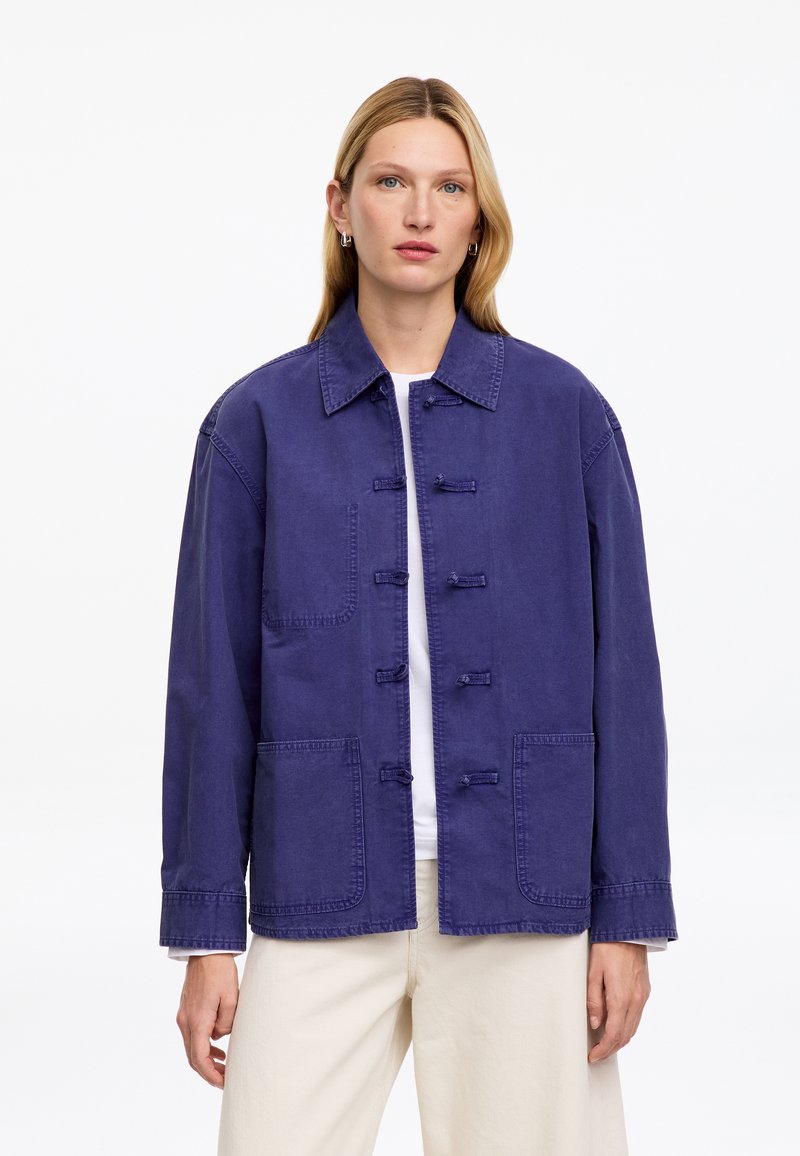 Femme aux longs cheveux blonds porte une veste ample bleu foncé avec de grandes poches avant, chemise blanche et pantalon beige clair, debout sur un fond blanc.