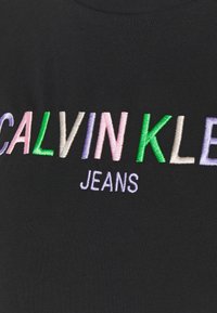 Černé bavlněné tričko s vícero barevně vyšívaným textem: "CALVIN KLEIN" v zelené, růžové, fialové a bílé barvě, s "JEANS" v bílé barvě níže.