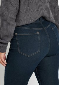 Jeans in denim blu scuro con un design aderente, caratterizzati da due tasche posteriori e cuciture marroni. Abbinati a un maglione a maglia grigia con texture.
