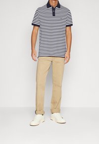 Muž v námořnickém a bílém pruhovaném polo triku, béžových rolled-up kalhotách a bílých teniskách stojící na neutrálním pozadí.