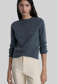 Grauer Wollpullover mit Rundhalsausschnitt, langen Ärmeln und gerippten Bündchen. Getragen über beigen Hosen, die eine weiche Textur und ein minimalistisches Design aufweisen.