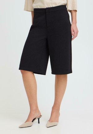 Frau trägt schwarze, knielange, maßgeschneiderte Shorts, beige Bluse und beigefarbene spitze Slingback-Absätze, steht vor einem einfarbigen Hintergrund.