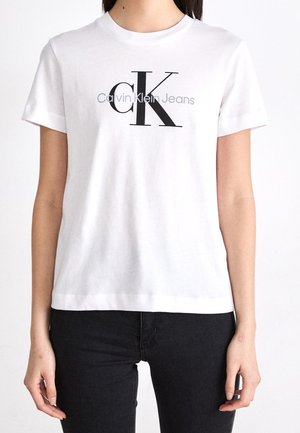 Bílé tričko s krátkým rukávem s černým logem "cK" a šedým logem "Calvin Klein Jeans" nošené s černými kalhotami.