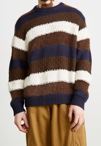 Stickad tröja med horisontella ränder i brunt, marinblått och krämvitt. Sticksig textur, ribbade ärmslut och nederkant, med ett grovt stickmönster.