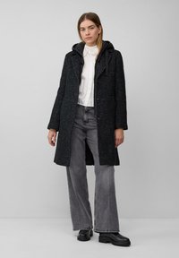 Vrouw in een zwart gevlekt jasje met capuchon over een wit shirt, brede grijze jeans en zwarte laarzen, staande tegen een effen lichtgrijze achtergrond.