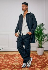 Schwarzer Knopf-über-Knopf-Mantel über einem hellbeigen Hemd, kombiniert mit dunklen Jeans und schwarz-weißen Sneakers, die ein dezentes Muster und Textur aufweisen.