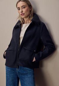 Giacca blu navy con colletto testurizzato, zip frontale e due tasche sul petto. Realizzata in un materiale morbido e strutturato, abbinata a jeans in denim blu.