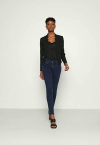 Casaco preto ajustado, top preto com renda, jeans skinny azul escuro e sandálias de salto pretas. A modelo está de pé contra um fundo neutro.