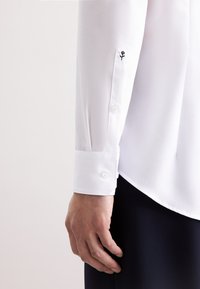 Weißes Langarmhemd mit geknöpften Manschetten, das ein kleines besticktes Rosendetail aufweist. Glattes Gewebe, klassisches Design und dezenter Glanz.