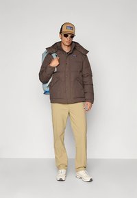 Brown puffer jacket with a hood, two front pockets, paired with beige pants and a tan cap. White sneakers and blue backpack are included.