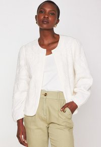 Donna che indossa un giubbotto bianco effetto tessuto sopra una maglietta bianca e pantaloni beige a vita alta con tasche con bottone, in piedi contro uno sfondo semplice.