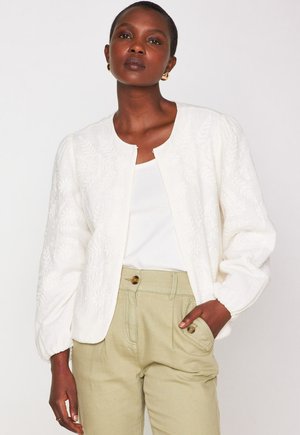 Donna che indossa un giubbotto bianco effetto tessuto sopra una maglietta bianca e pantaloni beige a vita alta con tasche con bottone, in piedi contro uno sfondo semplice.
