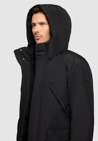 Black insulated jacket featuring a large hood, snap buttons, and a zippered pocket on the side. Smooth texture with a minimal design.