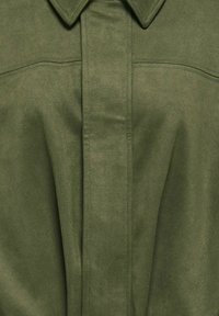 Košeľa z oliwkovo zelenej semišovej kože s ostrým golierom, skrytou légou so zapínaním na gombíky a detailom vodorovného švu cez hruď.