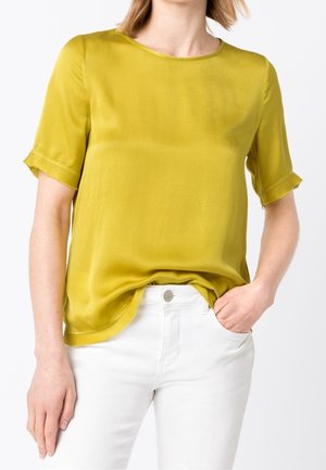 Vrouw met een mosterdgele korte mouwen blouse gestopt in een witte hoge taille broek met één hand in de zak.