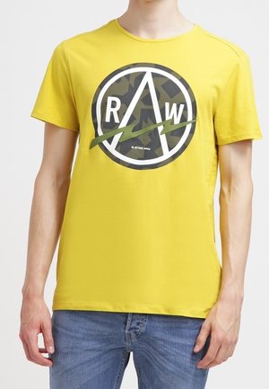 T-shirt en coton jaune avec un graphisme circulaire affichant "RAW" et un motif camouflage, col rond et manches courtes.