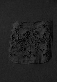 Sort stofskjorte med en dekorativ sort broderet lomme med blomster- og bladmønstre, der tilføjer teksturel kontrast.
