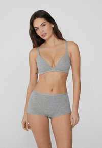 Joven mujer con cabello largo y oscuro, vistiendo un sujetador de algodón gris a juego y pantalones cortos tipo boyshort, de pie contra un fondo simple.