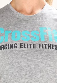 Camiseta de manga corta gris, hecha de tela suave. Presenta un texto turquesa en negrita "CrossFit" y "FORGING ELITE FITNESS" en negro debajo.