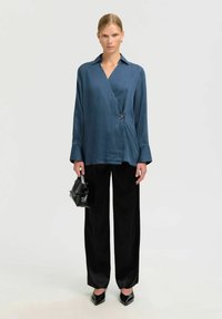 Camisa de satén azul con cuello, detallada con un cierre de botón lateral; combinada con pantalones de pierna ancha negros y un bolso negro.