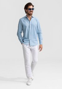 Lichtblauw overhemd met knopen, witte broek en witte sneakers. Model heeft bruin haar en draagt een donkere zonnebril. Glad stof met een casual pasvorm.