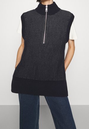 Veste sans manches en maille côtelée bleu marine avec demi-fermeture éclair argentée et col montant portée sur une chemise blanche à manches courtes et un jean bleu.