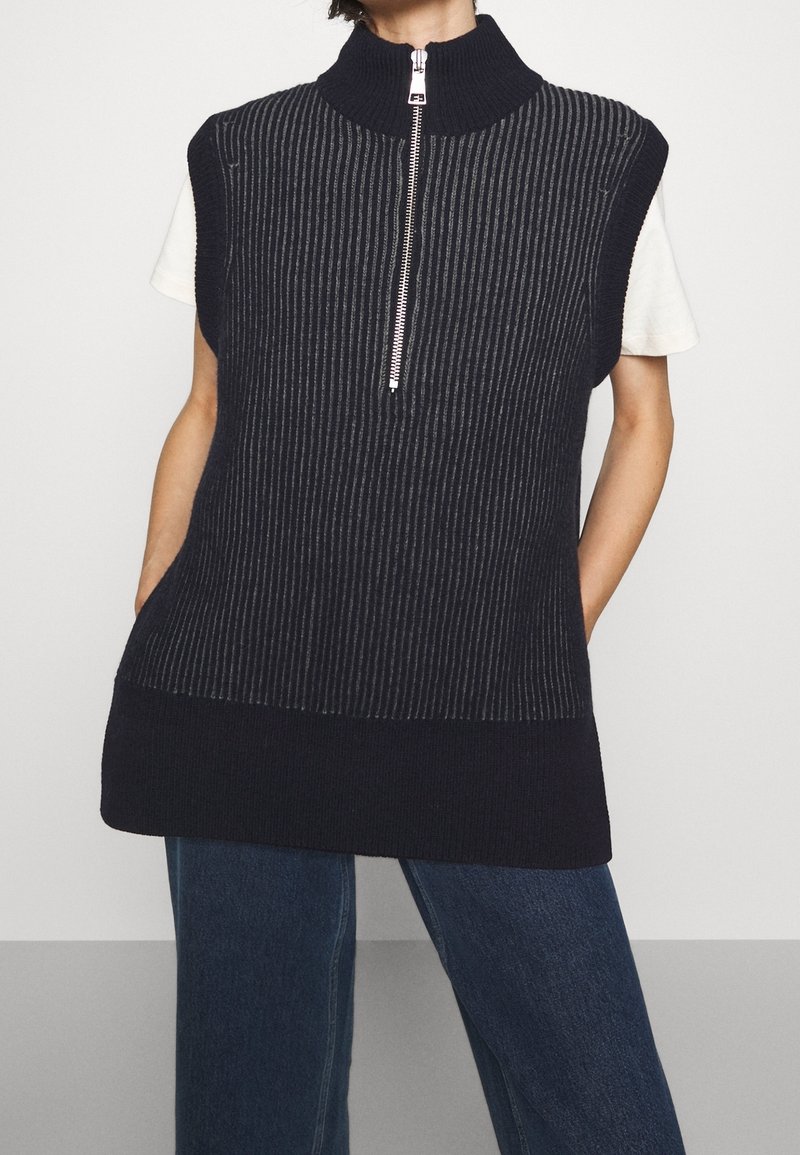 Veste sans manches en maille côtelée bleu marine avec demi-fermeture éclair argentée et col montant portée sur une chemise blanche à manches courtes et un jean bleu.