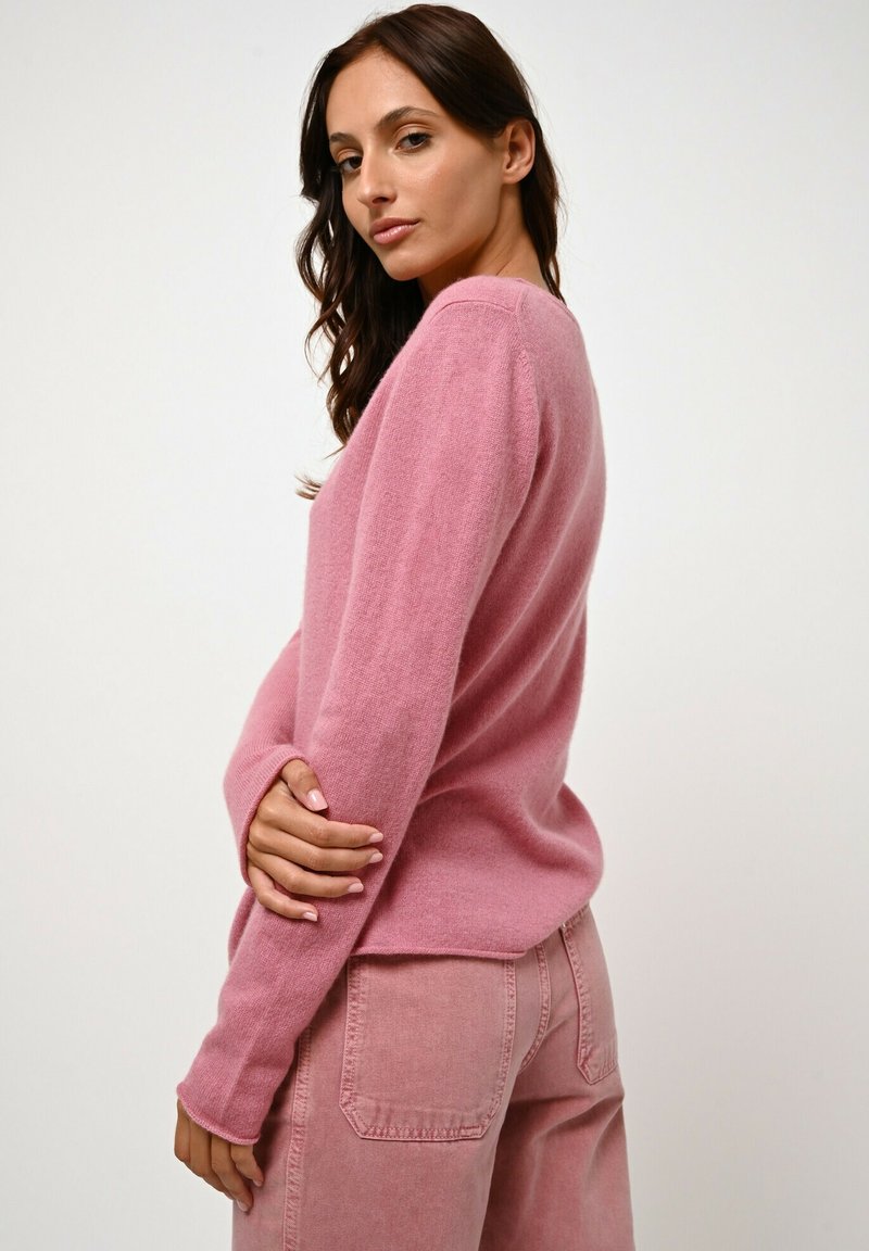 Rosa Strickpullover mit langen Ärmeln, rundem Halsausschnitt und lockerer Passform. Weicher Stoff, kombiniert mit passenden rosa Jeans mit Gesäßtaschen.