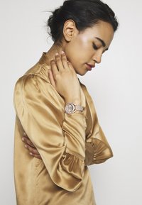 Guld satinblus med puffärmar, med en rund silverklocka med en beige rem och kristalldetaljer på boetten.