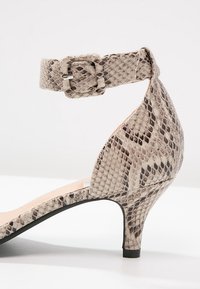 Chaussure à talon kitten beige et marron avec motif peau de serpent, bride de cheville et fermeture à boucle sur fond blanc.