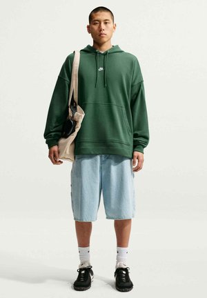 Joven con una sudadera verde de Nike con capucha, pantalones cortos azul claro hasta la rodilla, zapatillas negras, calcetines blancos y llevando un bolso beige al hombro.