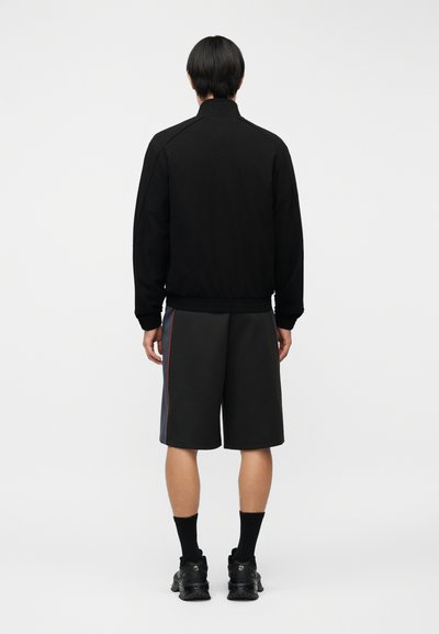 Άνδρας με την πλάτη γυρισμένη, φοράει μαύρο μπουφάν, μαύρο και γκρι μακρύ σορτς με κόκκινη ρίγα, μαύρες κάλτσες και μαύρα αθλητικά παπούτσια σε λευκό φόντο.