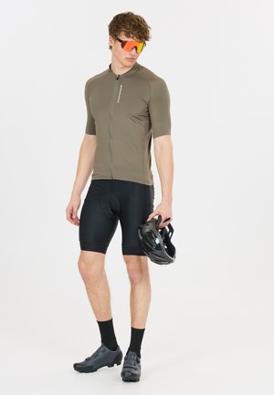 Mann in olivgrünem Radtrikot, schwarzen gepolsterten Shorts, Sonnenbrille und Schuhen hält einen schwarzen Fahrradhelm vor weißem Hintergrund.