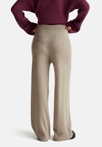 Béžové pletené kalhoty s širokými nohavicemi, s žebrovaným pasem a manžetami, zobrazené zezadu, zdůrazňující hladkou texturu a elegantní design.