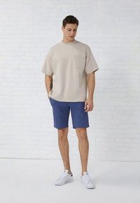 Beiges Baumwoll-T-Shirt mit kurzen Ärmeln und einer Tasche, kombiniert mit blauen Shorts. Weiße Turnschuhe; vor einem weißen Ziegelstein-Hintergrund.
