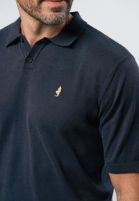 Vyras dėvi tamsiai mėlyną trumpomis rankovėmis polo marškinėlį su subtiliu smėlio spalvos siuvinėtu logotipu kairėje krūtinės pusėje, matomas apatinis veidas ir kaklo ūsai.