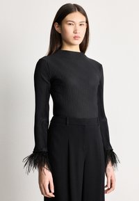 Žena v čiernom rebrovanom tričku s dlhými rukávmi ukončenými perím a v čiernych nohaviciach s vysokým pásom, stojaca pred obyčajným pozadím.