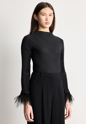 Žena v čiernom rebrovanom tričku s dlhými rukávmi ukončenými perím a v čiernych nohaviciach s vysokým pásom, stojaca pred obyčajným pozadím.