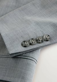 Hellgrauer Anzugärmel mit vier dunkelgrauen Knöpfen in der Nähe der Manschette, die feine, strukturierte Stoffe und eingenähte Knopflöcher zeigt.