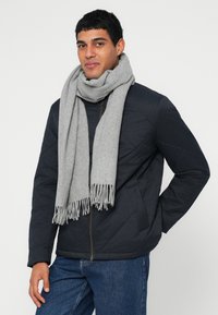 Casaco acolchoado azul-marinho com fecho de correr à frente e bolsos laterais, combinado com um cachecol longo cinza claro com franjas. A modelo usa jeans escuros.