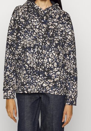 Person, der einen schwarzen Kapuzenpullover mit beige- und blauem abstraktem Farbspritzmuster und einer dunkelblauen Jeans trägt.