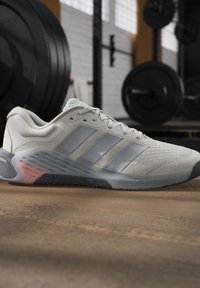 Светлосив Adidas маратонка с три ивици, почиваща на пода в залата, с размазани тежести и фитнес оборудване на заден план.