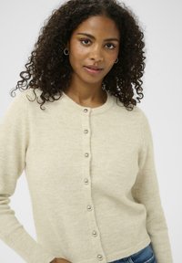 Hellbeige Strickjacke aus weichem, strukturiertem Stoff. Sie hat einen runden Ausschnitt und sieben metallische Knöpfe auf der Vorderseite. Lange Ärmel.