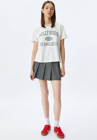 Camiseta de algodón blanca con gráfico de "HOLLYWOOD", combinada con una falda gris con pliegues. La modelo lleva zapatillas rojas con calcetines blancos. Look suave y casual.