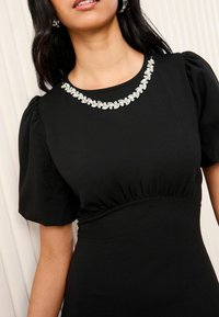 Zwarte jurk met opbollende mouwen en een geplooide taille. Beschikt over een decoratieve kristallen versiering langs de ronde halslijn.