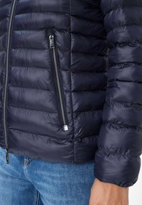 Marineblaue Steppjacke mit ge quilteter Textur, ausgestattet mit einem Reißverschluss mit metallischen Akzenten und elastischen Bündchen. Sichtbare Nähte betonen das Design.
