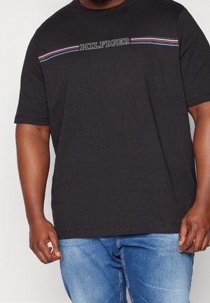 Homme portant un T-shirt noir avec le texte "HILFIGER" et des bandes rouges, blanches et bleues sur la poitrine, associé à un jean bleu.