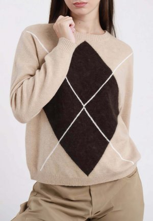 Donna che indossa un maglione beige con un grande motivo a rombi marrone scuro e linee diagonali bianche, abbinato a pantaloni beige.