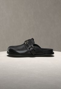 Μαύρο δερμάτινο παπούτσι mule με στρογγυλεμένο δάχτυλο, λεπτομέρεια με λουράκι και πόρπη, και χαμηλή επίπεδη σόλα σε φωτεινή επιφάνεια.