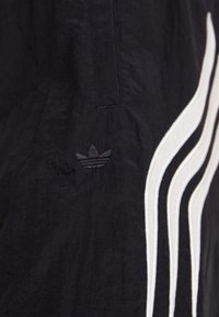Joggers de tela negra con un logo bordado y tres franjas blancas a los lados, superficie texturizada con costuras visibles.